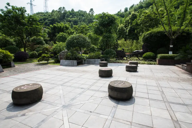 A serene garden featuring five round stone stools on a paved area, surrounded by lush greenery and trees under a clear sky.